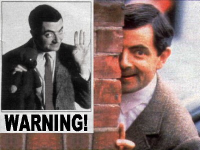 Rowan Atkinson, als Mr. Bean, kijkt rechtsom de hoek van een muur lachend naar ons. Op de muur hangt een zwart-witte poster met een foto van Mr. Bean erop, boven de tekst 'Warning!'.