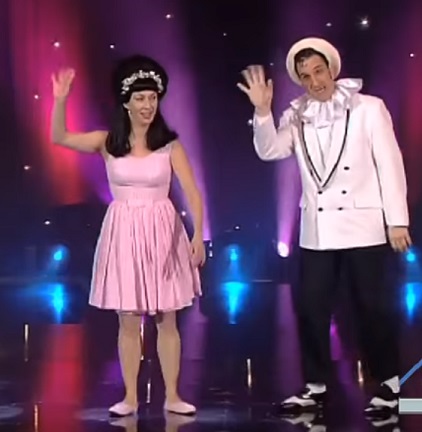 Corinne (links) en Gilles (rechts), als Shirley en Dino, zwaaien naar het publiek vanop het podium. Shirley draagt een kort, roze jurkje en heeft een opvallend hoog kapsel met diadeem; Dino draagt een zwart/wit kostuum met pierrotkraag en een witte pet.