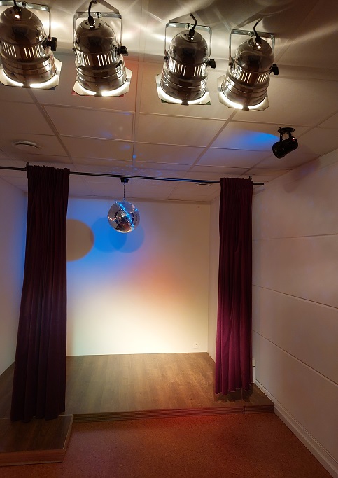 Een klein podium wordt verlicht door vier schijnwerpers. Boven het podium hangt een discobal, beschenen door blauw licht, wat blauwe lichtpuntjes reflecteert op de muren en het plafond.
