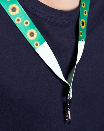 Een Sunflower-hanger hangt om de hals. De hanger heeft een groen 'snoer' waarop zonnebloemen afgebeeld staan. Onderaan is de hanger wit. Een sleutel hangt aan de hanger.