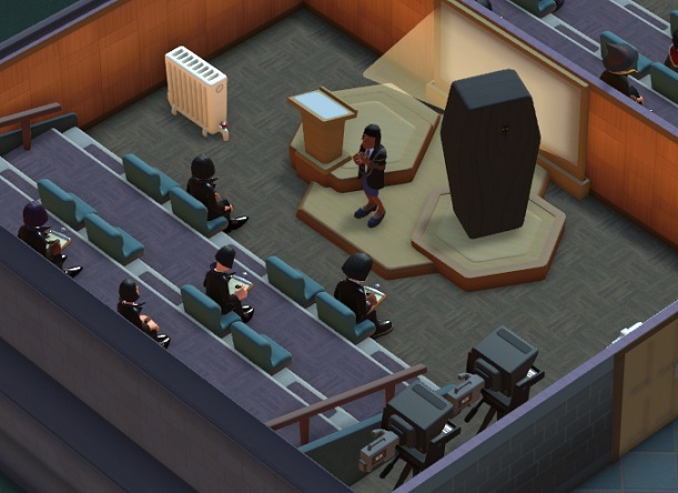 Vogelperspectief van een Lecture Theatre waar net een Dark Art-les plaatsvindt. De Docente staat midden bovenaan; rechts van haar staat een grote, zwarte doodskist. Links van de Docente staat een grote Radiator. Rechtsonder staan twee Autocues. Vijf Goth-Studenten wonen de les bij.