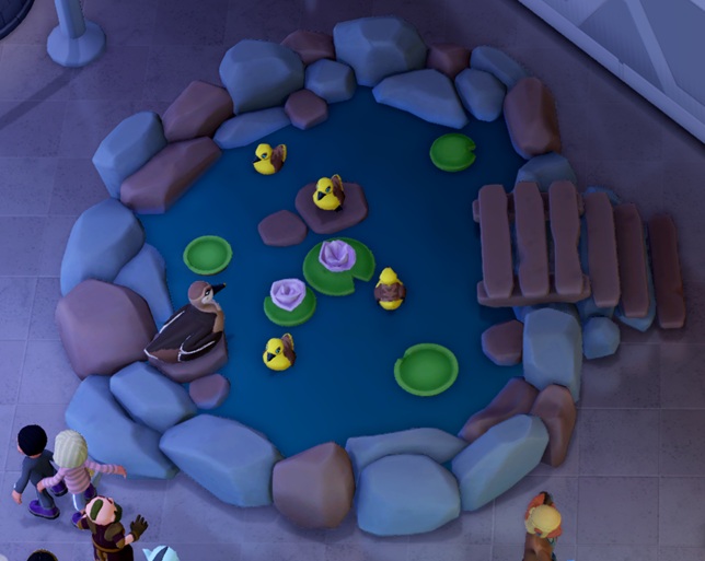 Vogelperspectief van een Duck Pond Diorama: een rond Eendenvijvertje met waterlelies en vier rubberen eendjes op het water. Links zit een volwassen Eend het vijvertje te overzien. Rechts loopt een houten bruggetje over de stenen rondom de vijver. Bezoekers lopen linksonder en rechtsonder voorbij.